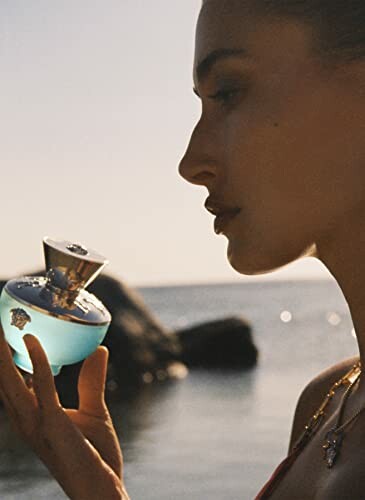 Woman holding a blue perfume bottle near a body of water.